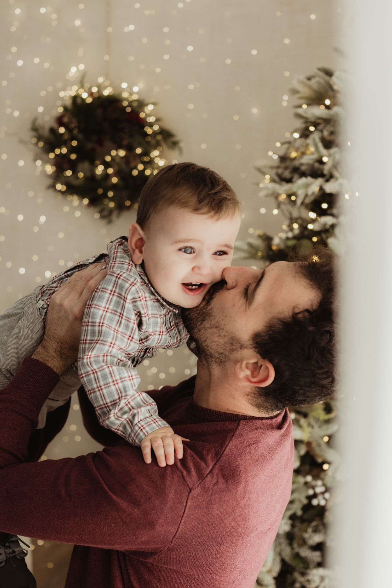 Playful moment between parent and baby near decorated Christmas tree with twinkling lights.