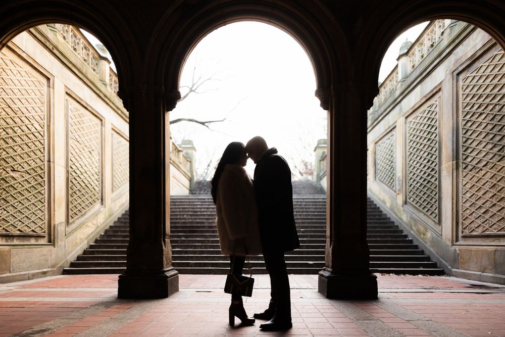 Two silhouettes stand embracing in an archway at Central Park's Bethesda Terrace with stone staircases flanking them.