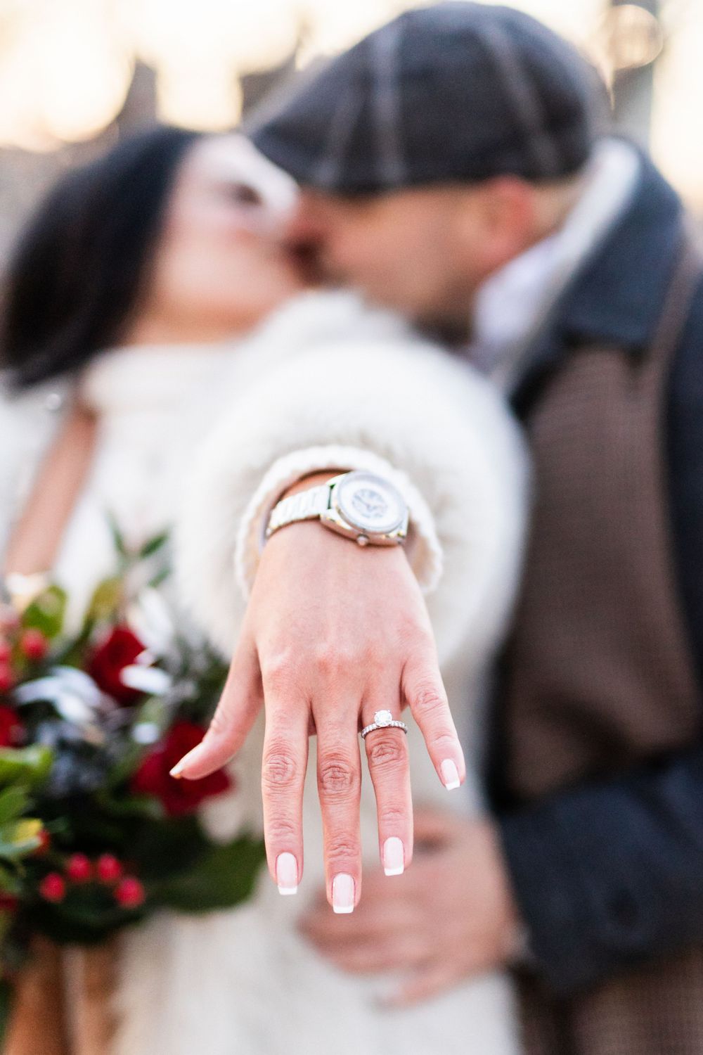 Close-up of an engagement ring with manicured nails and wedding flowers in soft focus.
