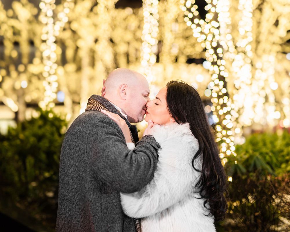 A couple shares a romantic kiss outdoors with twinkling string lights creating a magical backdrop in the evening.