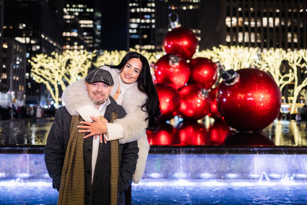 A couple poses by festive holiday decorations with giant red ornaments and twinkling lights.