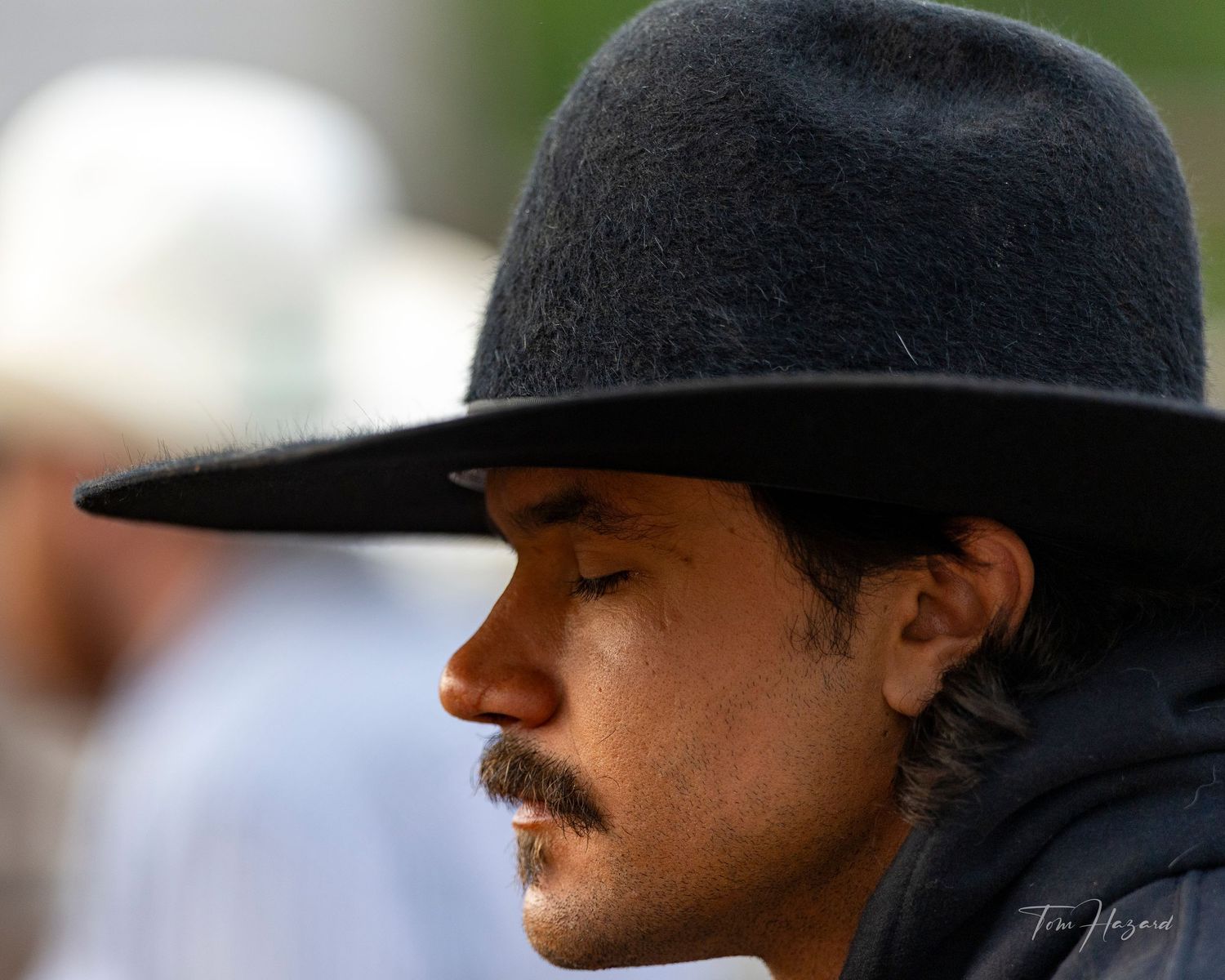 A young man with a mustache wearing a black wide-brim cowboy hat, photographed in close-up profile view outdoors.