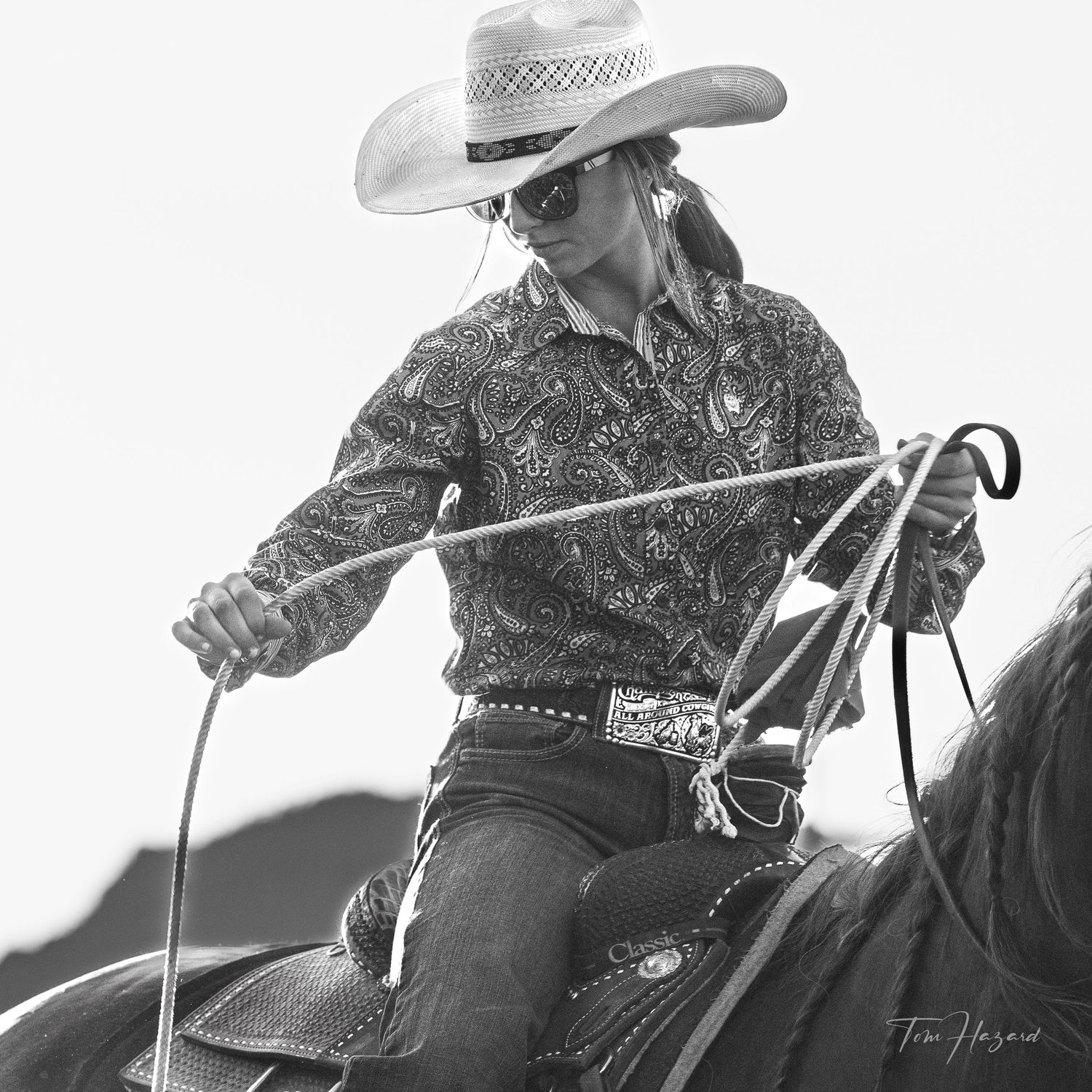 Black and white photo of a cowgirl in a patterned shirt and cowboy hat holding reins while riding a horse.