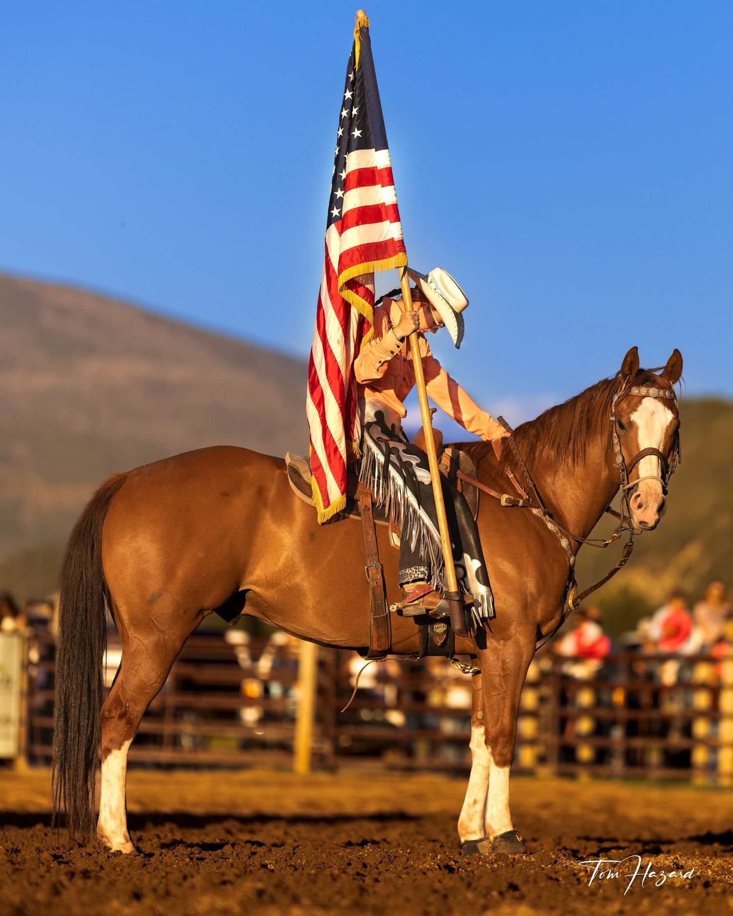 Rider on chestnut horse holding American flag at a rodeo arena with mountains in the background.