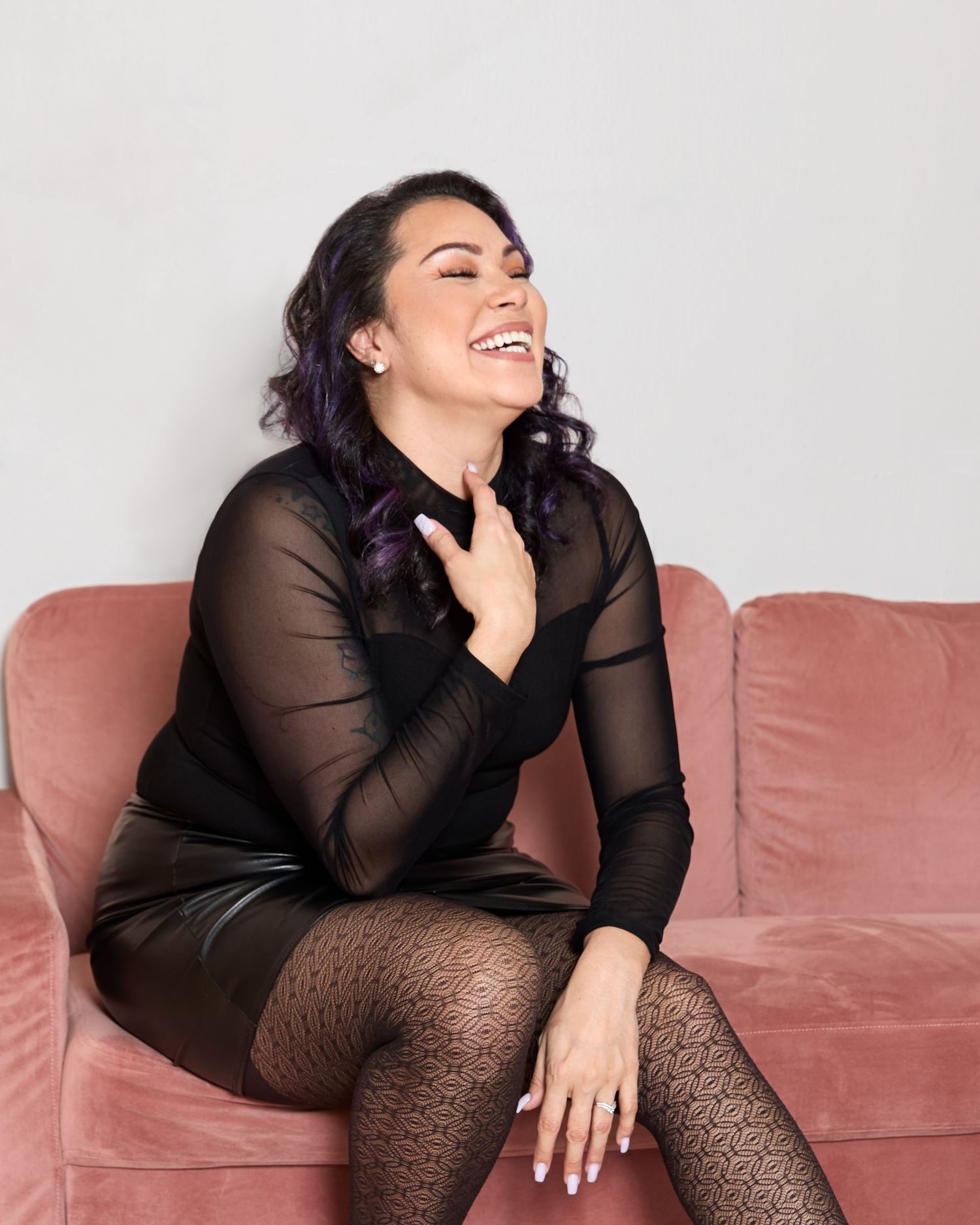 Joyful portrait on pink couch wearing sheer black dress and patterned tights.
