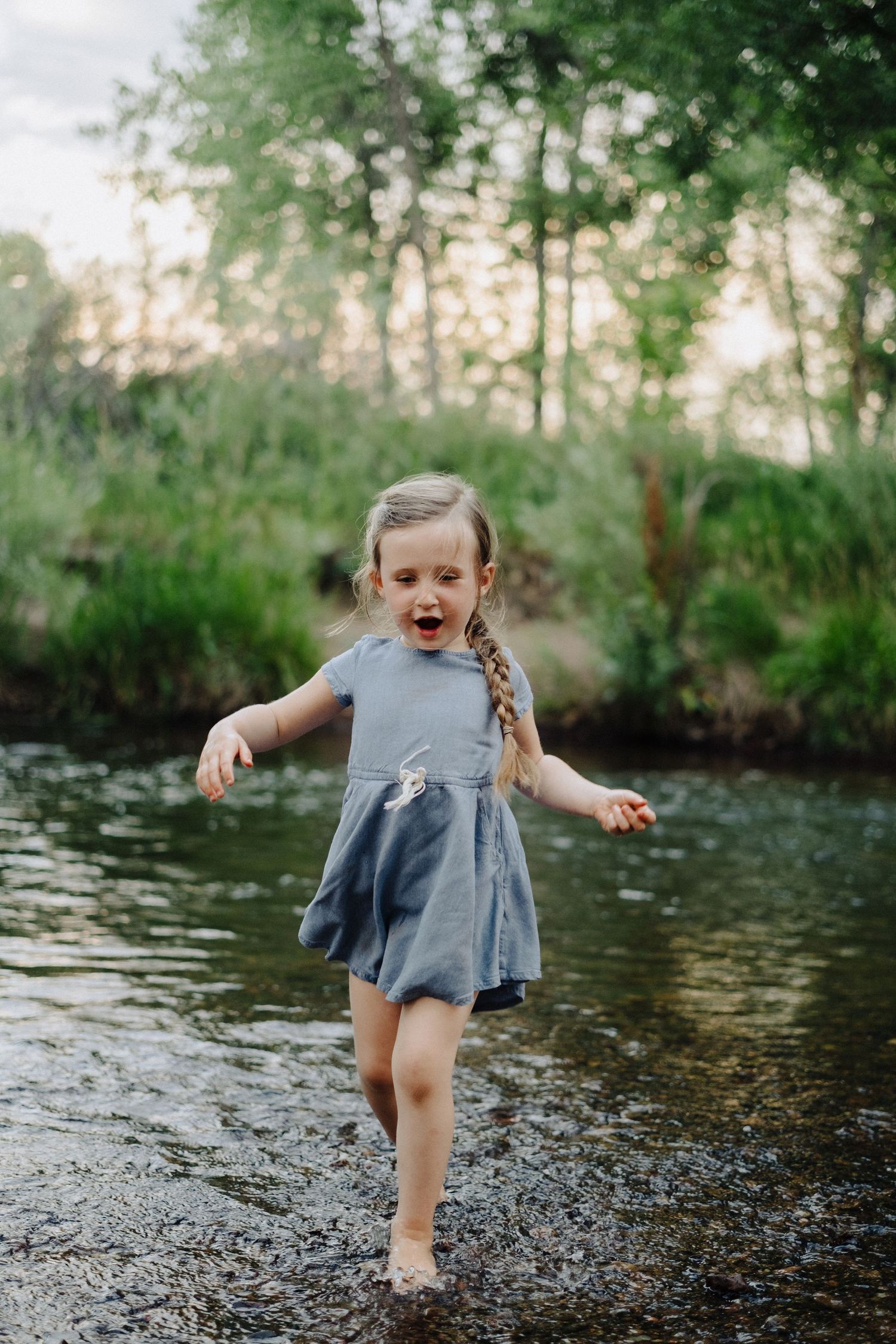 Young child in a blue dress playing and splashing in a creek on a sunny day.