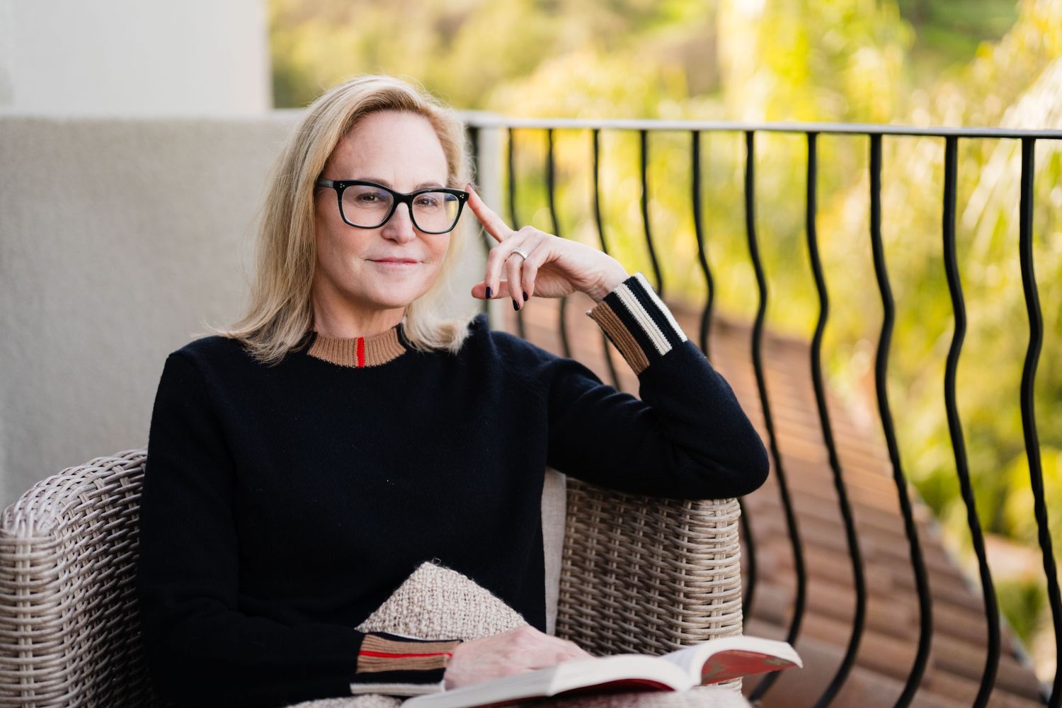 Person wearing glasses sits comfortably in a chair reading a book on a balcony.