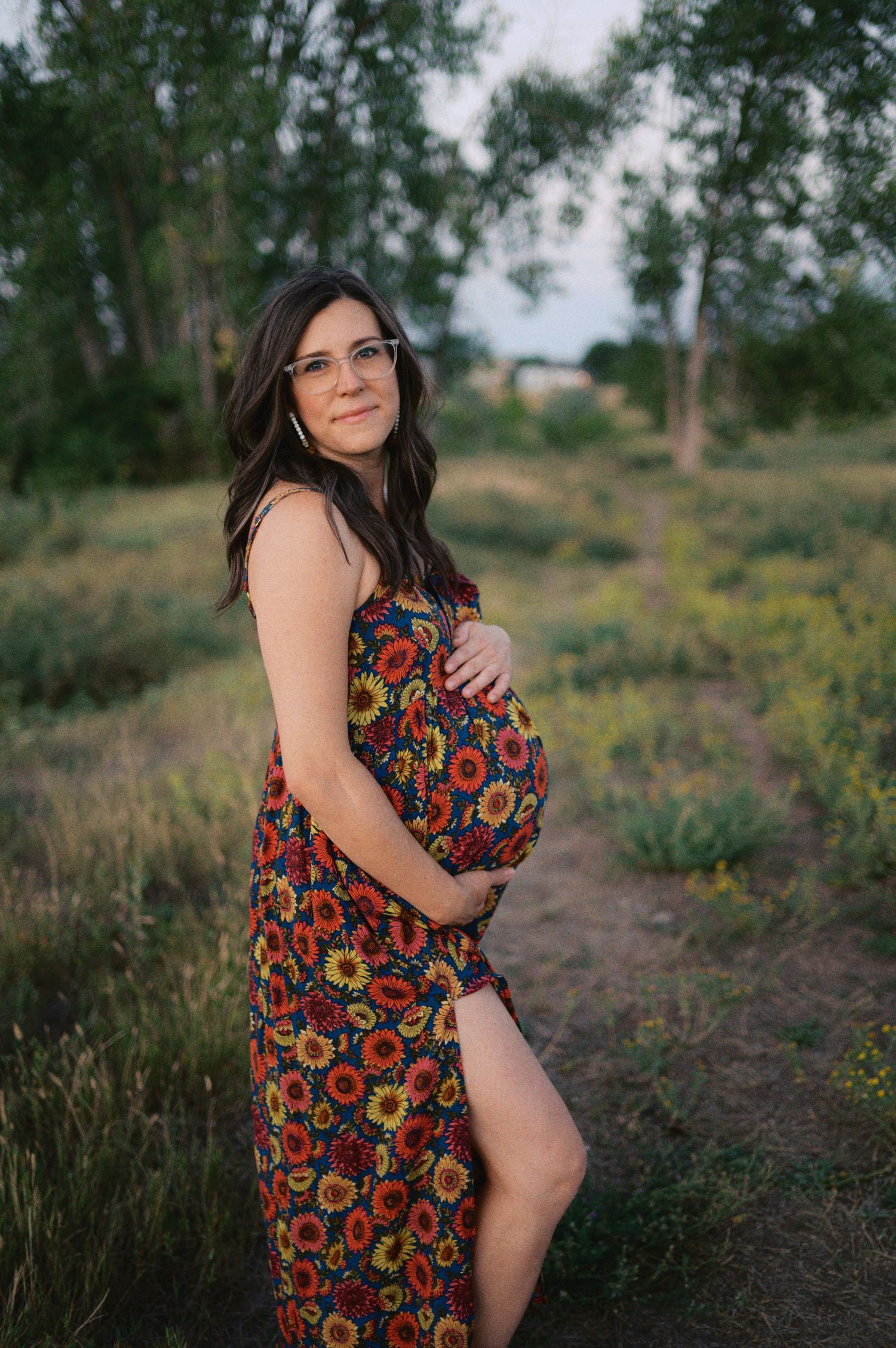Expecting mother in a colorful floral dress poses outdoors on a nature trail during sunset.