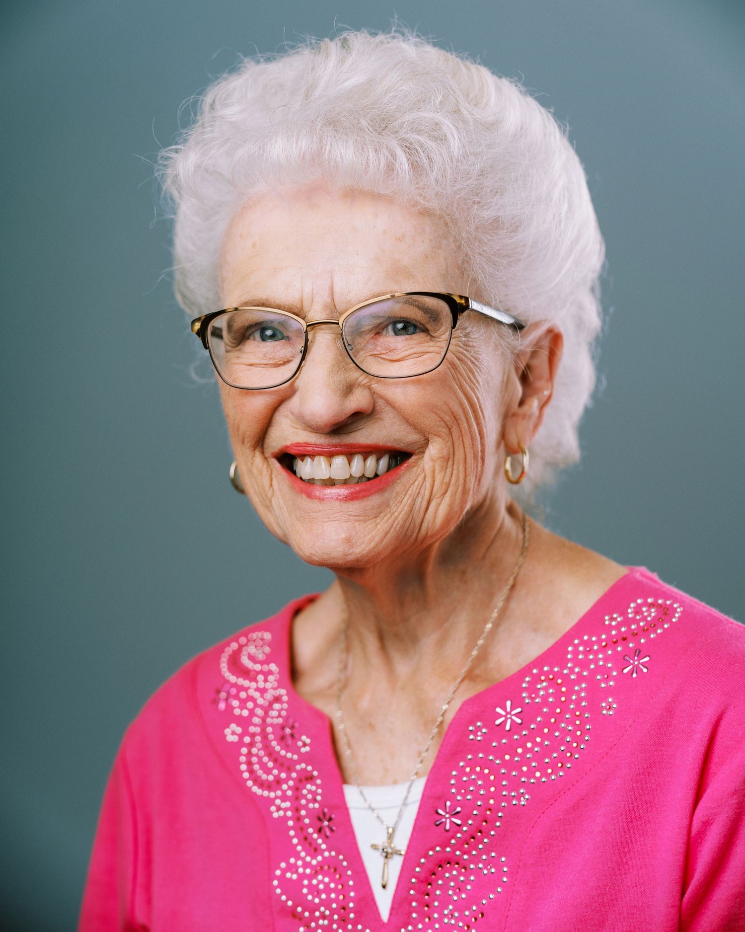 Senior in pink sequined top and glasses smiling warmly against blue backdrop.