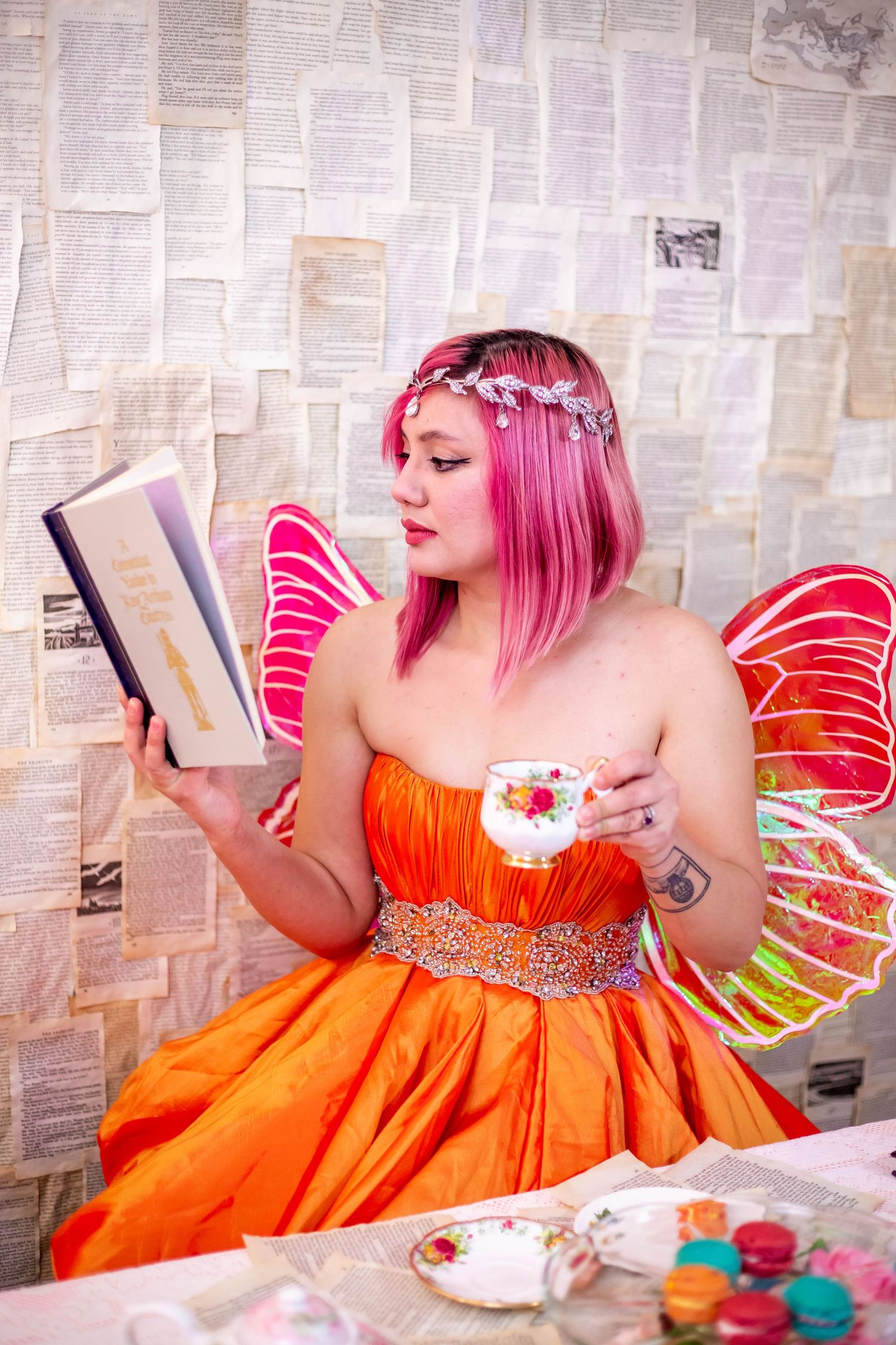 A whimsical fairy costume photoshoot featuring orange dress and pink wings against a paper backdrop.