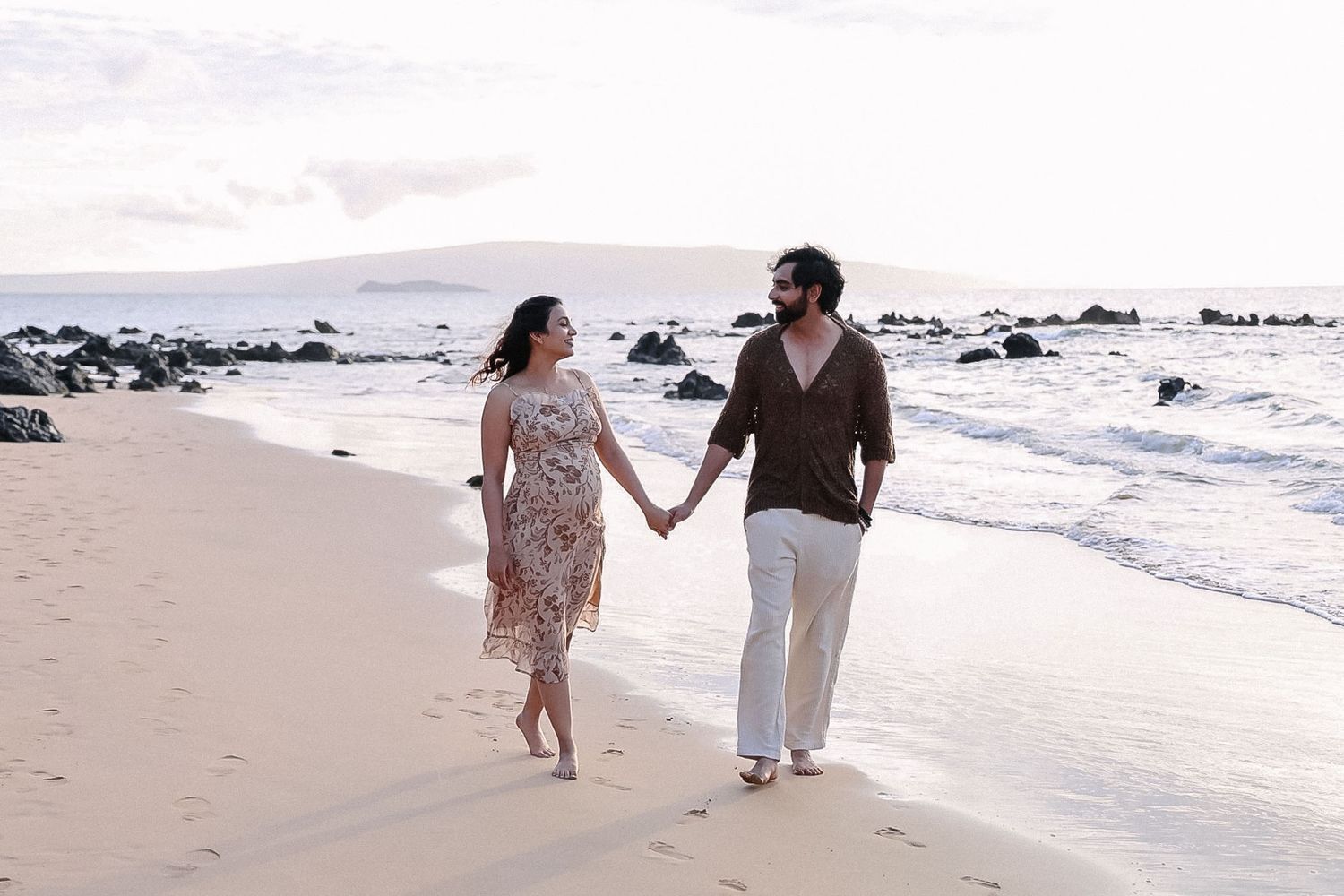 Couple walking hand in hand along a scenic beach shoreline during sunset with waves crashing nearby.