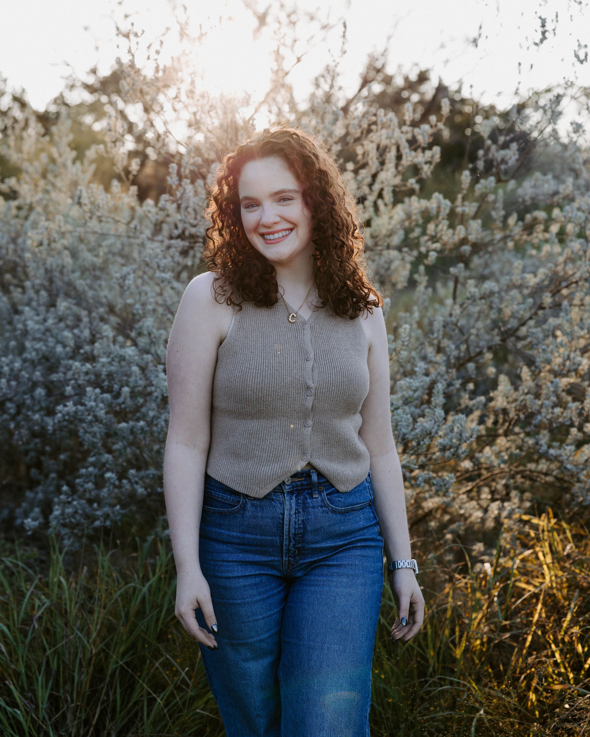 San Antonio Photographer Headshot. Person in beige sweater and jeans poses in front of blooming spring bushes at sunset.