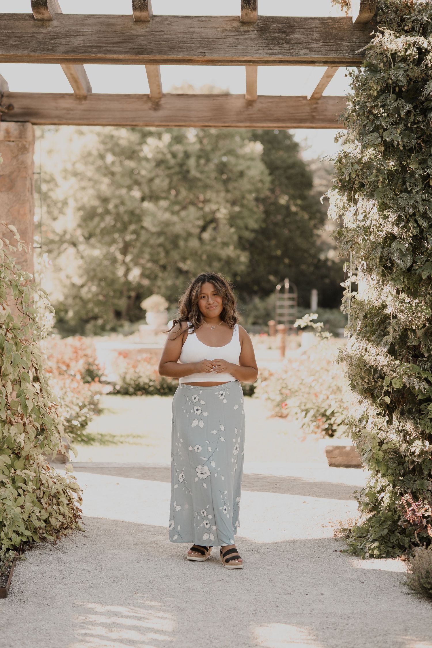 Person in white dress sitting on bench surrounded by lush garden greenery and wooden beams.