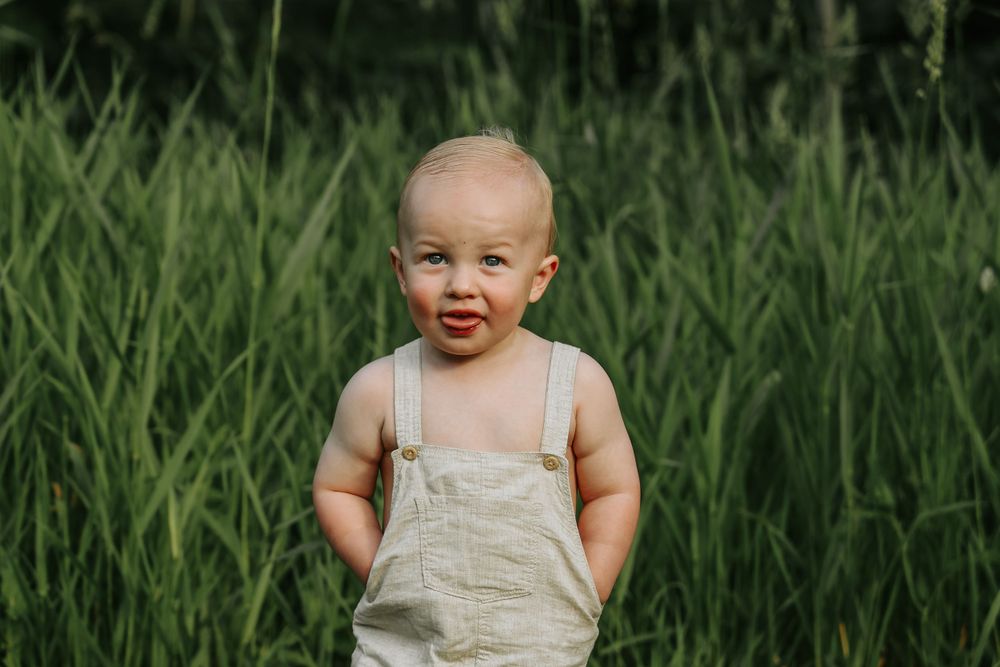 A young child in overalls smiles while standing in tall grass.