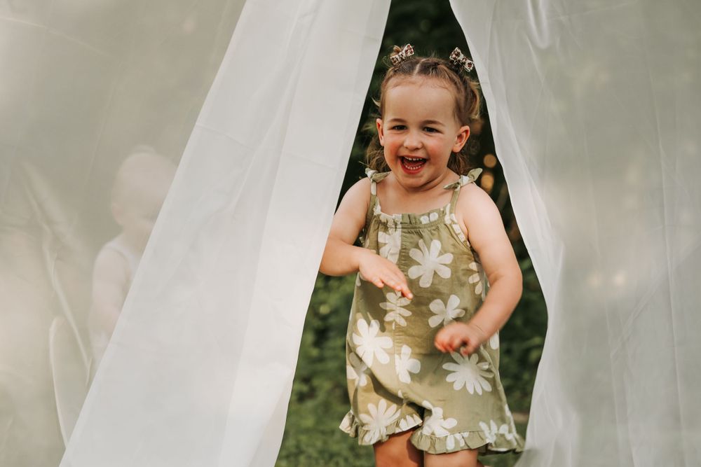 A child plays peek-a-boo through white curtains outdoors.