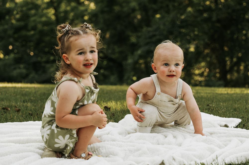 Two young children sit together on a white blanket in a sunny garden.
