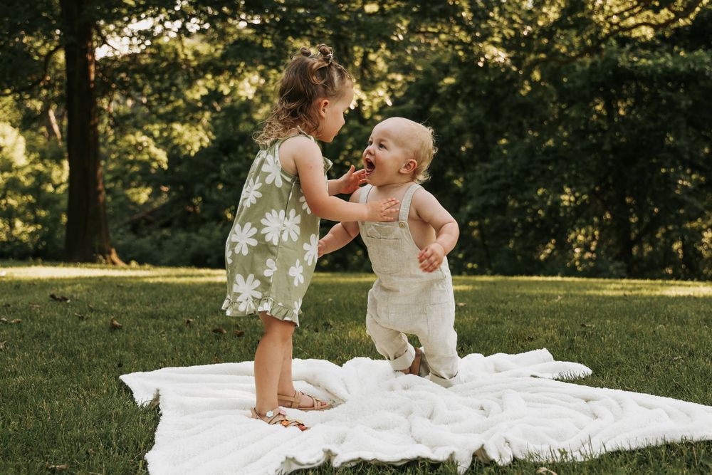 Two young children play together on a white blanket in a sunlit garden.