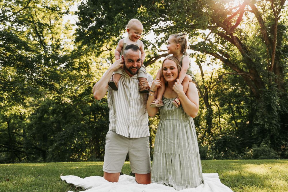 A family poses together on a white blanket in a sunny park setting.