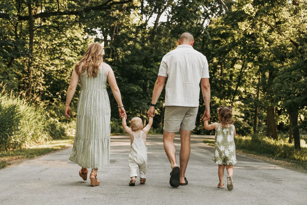 A family walks hand in hand down a sunlit path surrounded by trees.