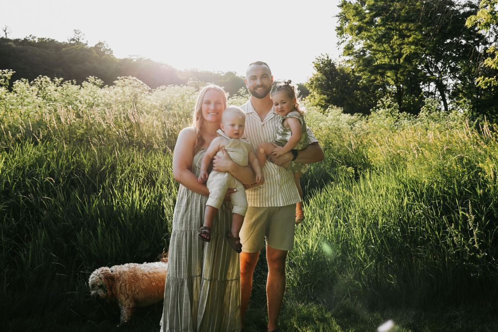 A family stands together in tall grass at sunset with their dog.