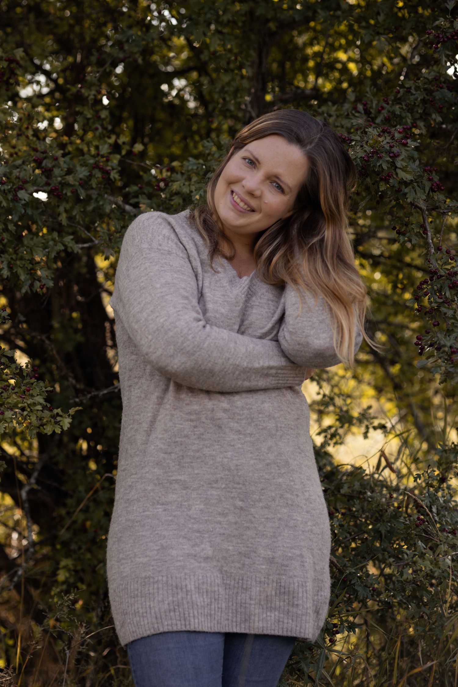 Person wearing a cozy gray sweater and jeans poses outdoors against autumn foliage background.