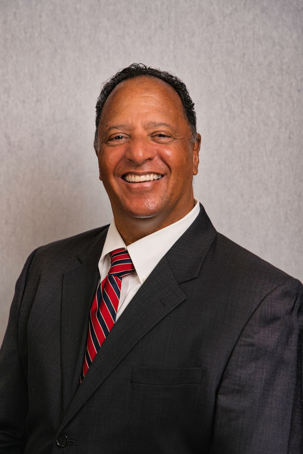 Professional headshot of man in dark suit with red striped tie against grey background.