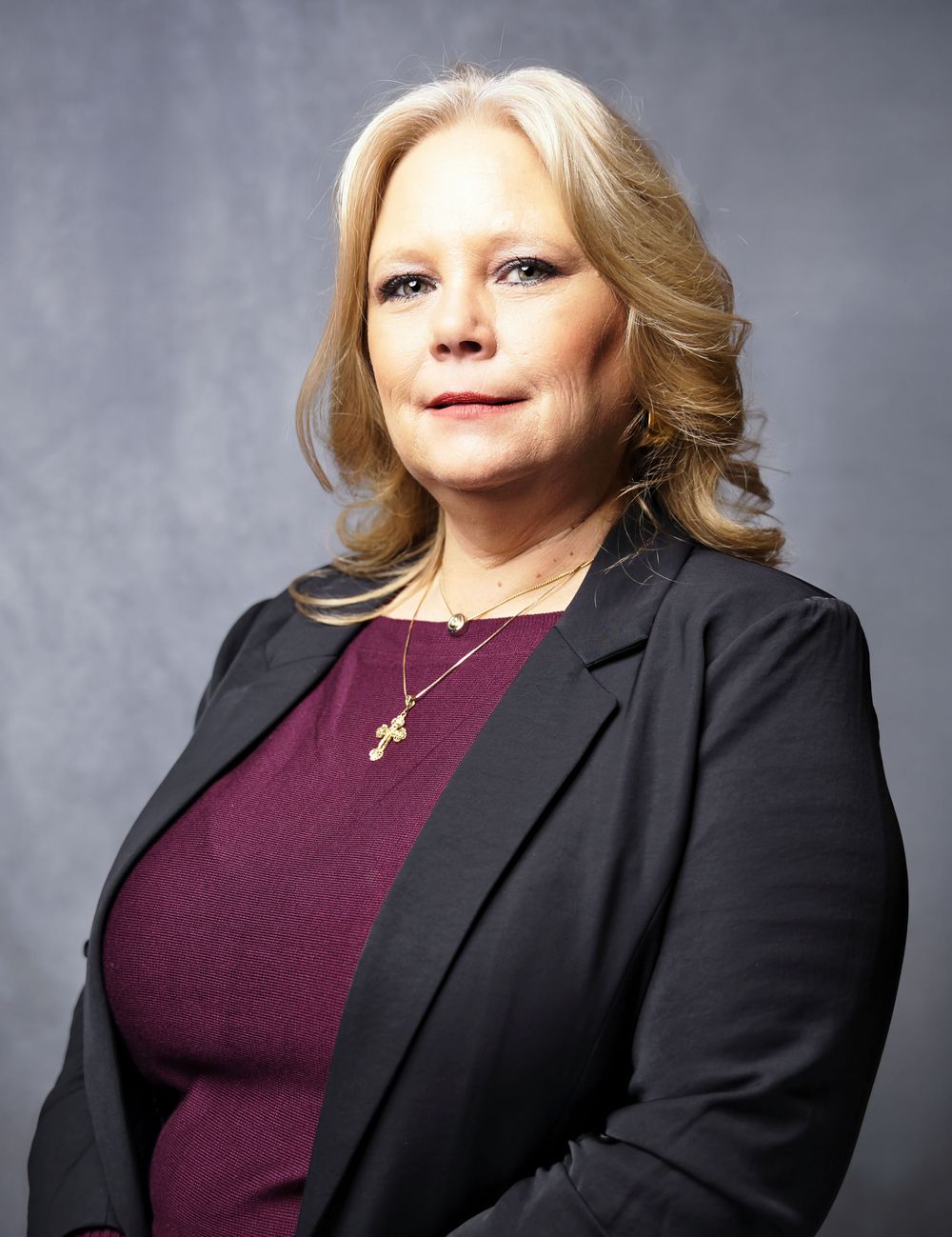 Professional studio portrait of someone wearing a black blazer and burgundy top with a cross necklace against a gray background.