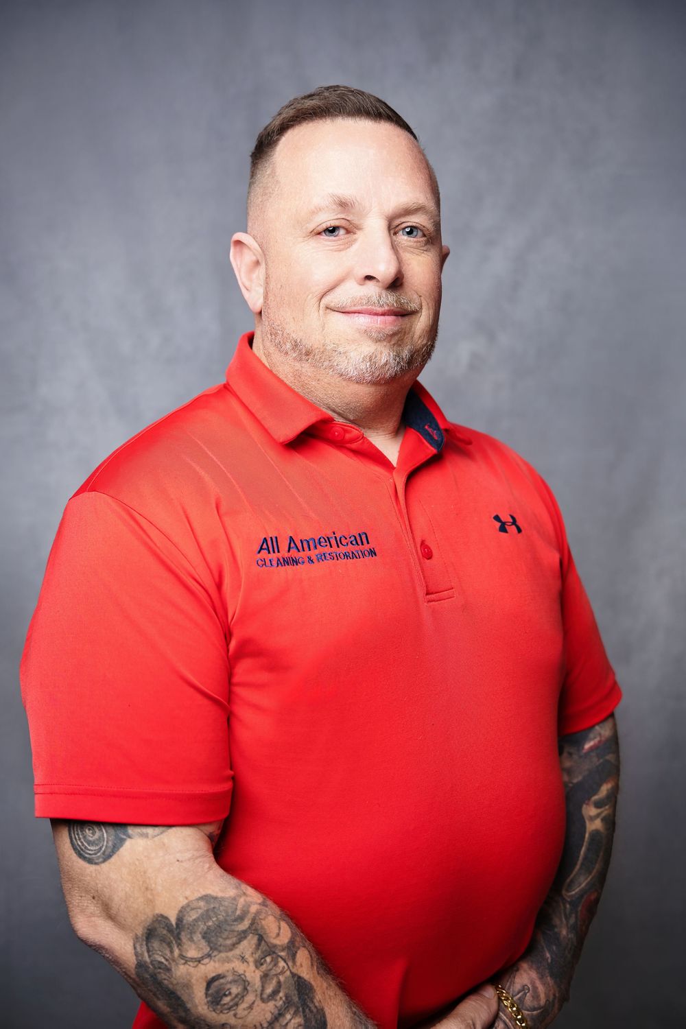 Professional portrait of someone in a red Under Armour polo shirt with visible arm tattoos against a gray background.