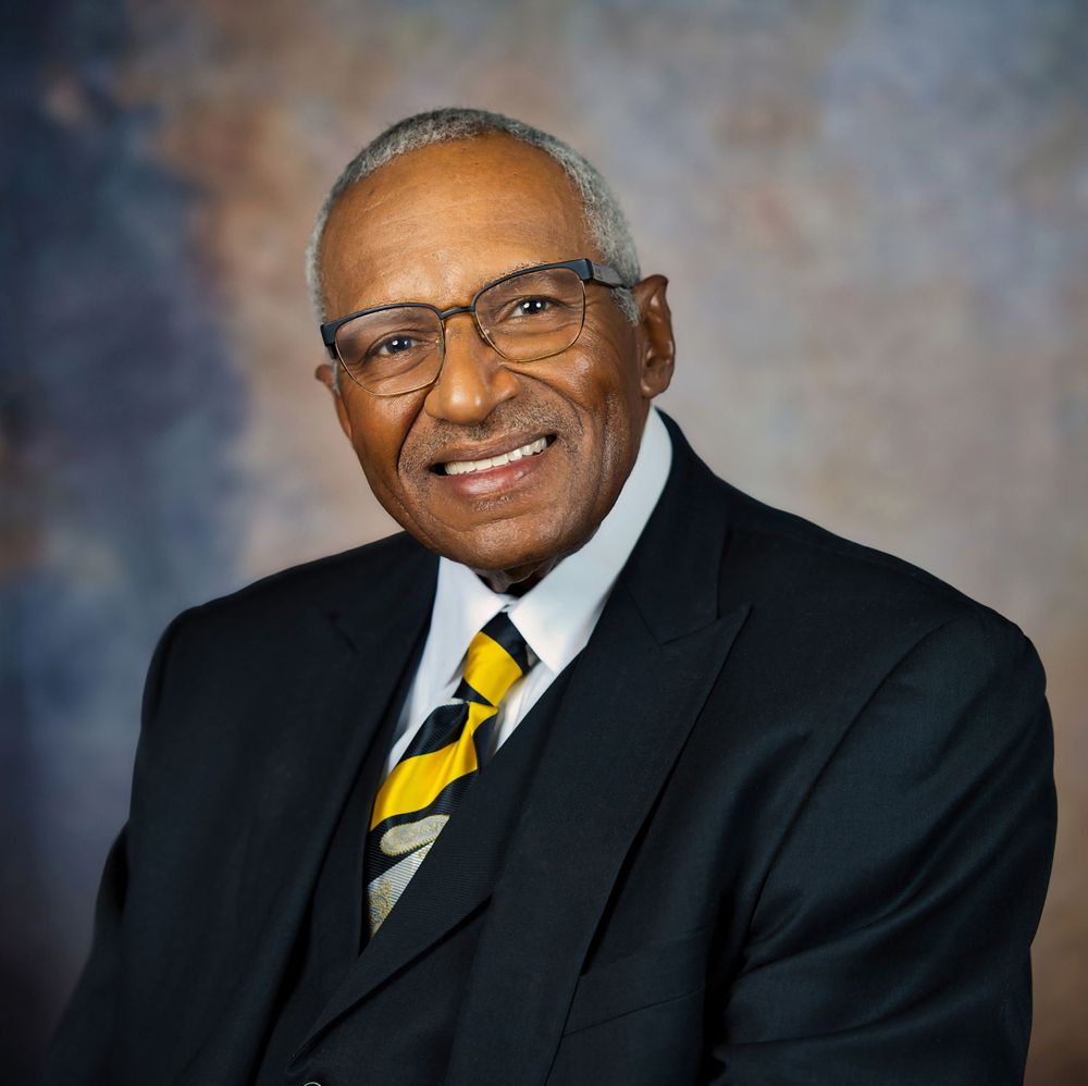 Professional headshot of a distinguished person wearing glasses and a yellow striped tie against a blue background.