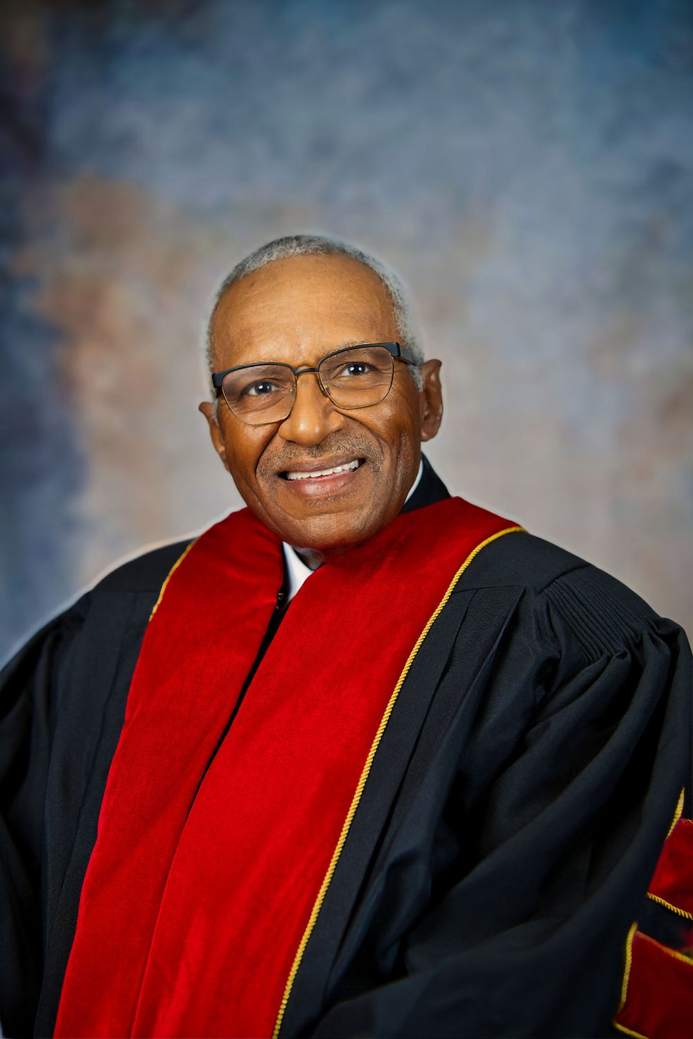 Professional portrait of a scholar wearing academic regalia with a red stole and black robe against a mottled background.