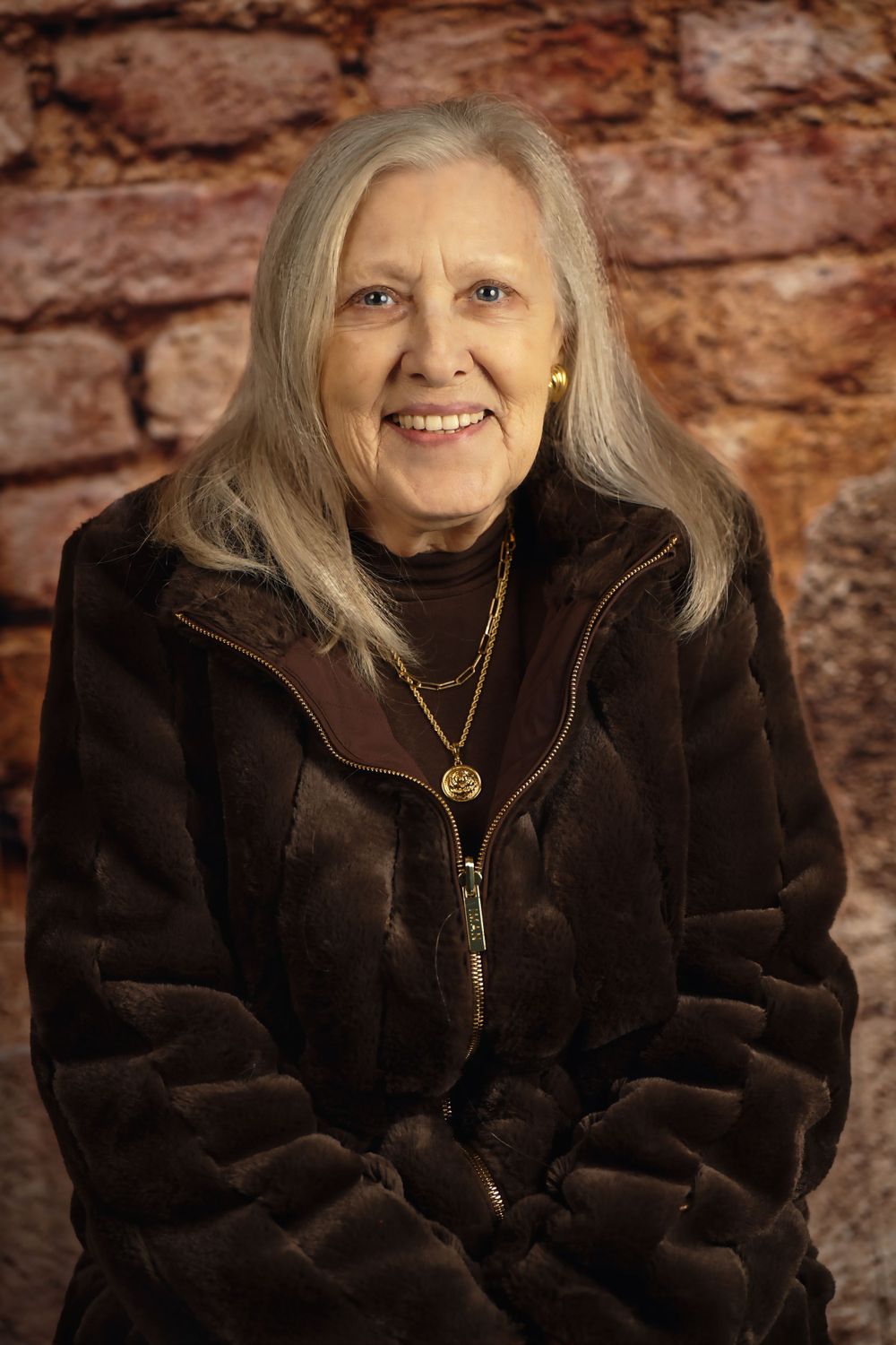 Portrait of older woman with long grey hair wearing brown fur coat against brick wall.