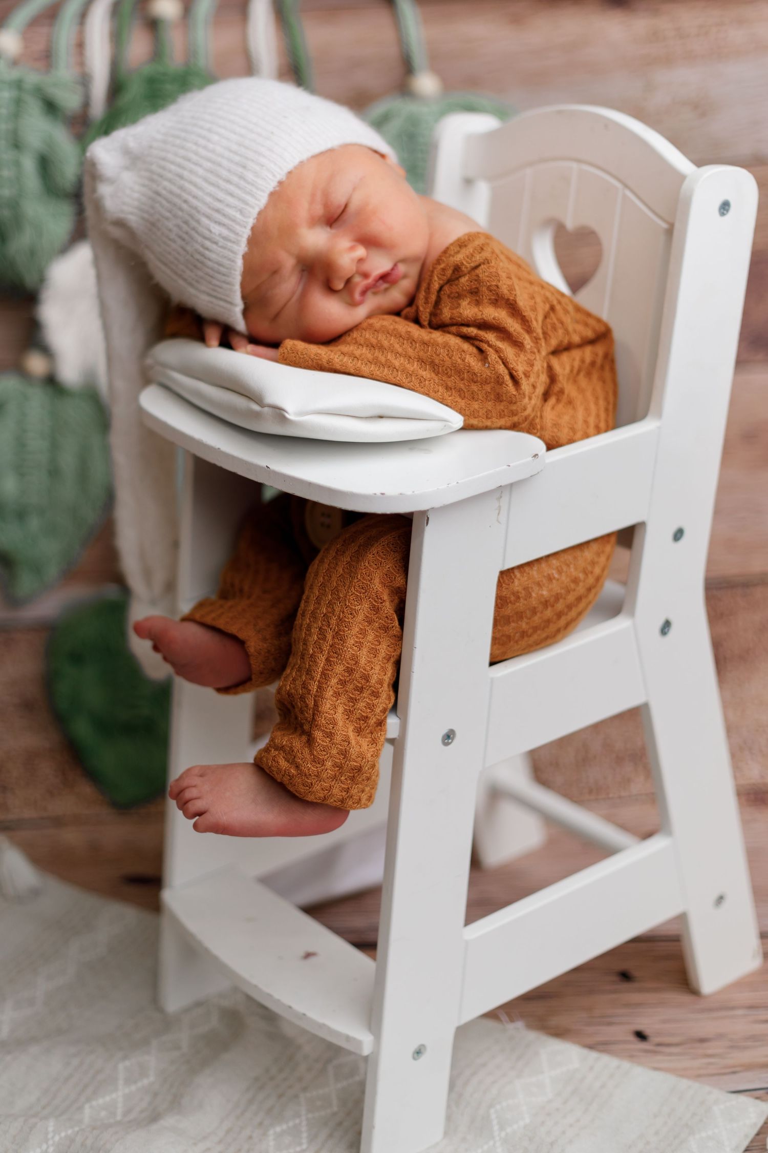 Sleeping baby wearing knit hat and orange outfit in miniature white wooden chair.