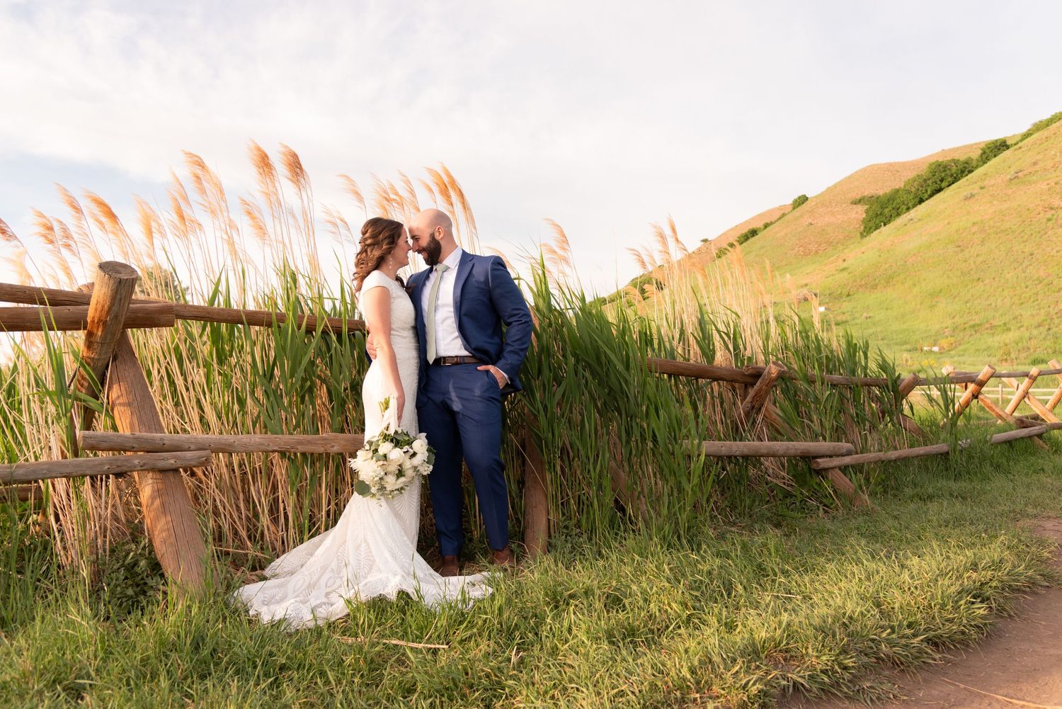 A couple embraces in a romantic moment on a hillside with tall grass at sunset.