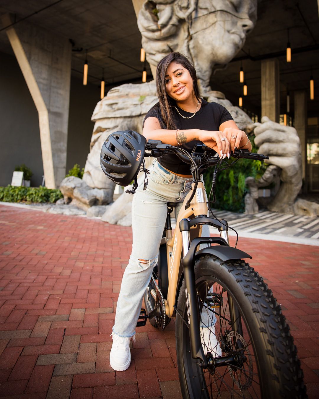 A person leans on a fat tire bicycle while smiling, with a decorative stone sculpture in the background.