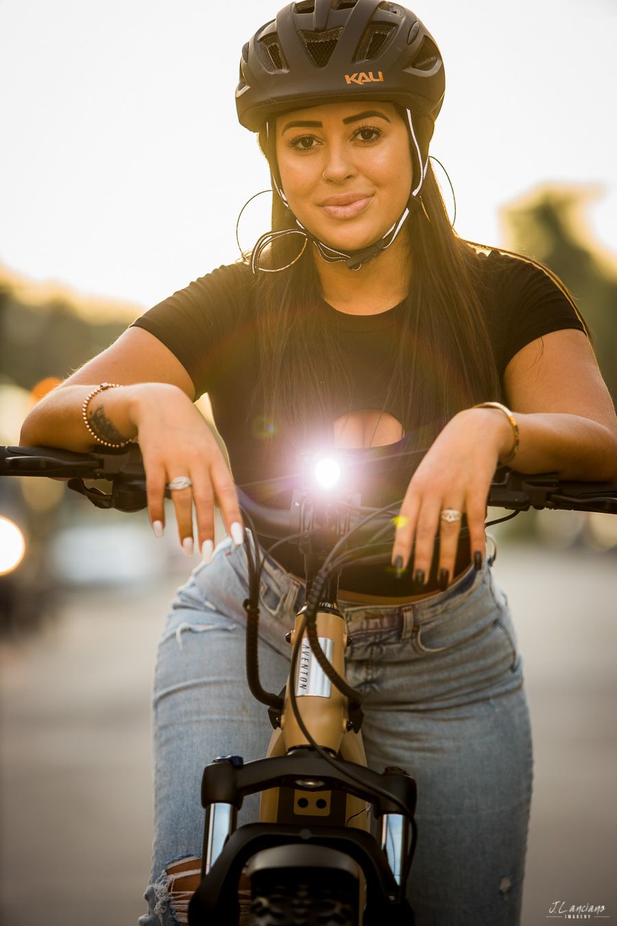 Cyclist wearing protective helmet and hoop earrings poses on bike at sunset with lens flare.