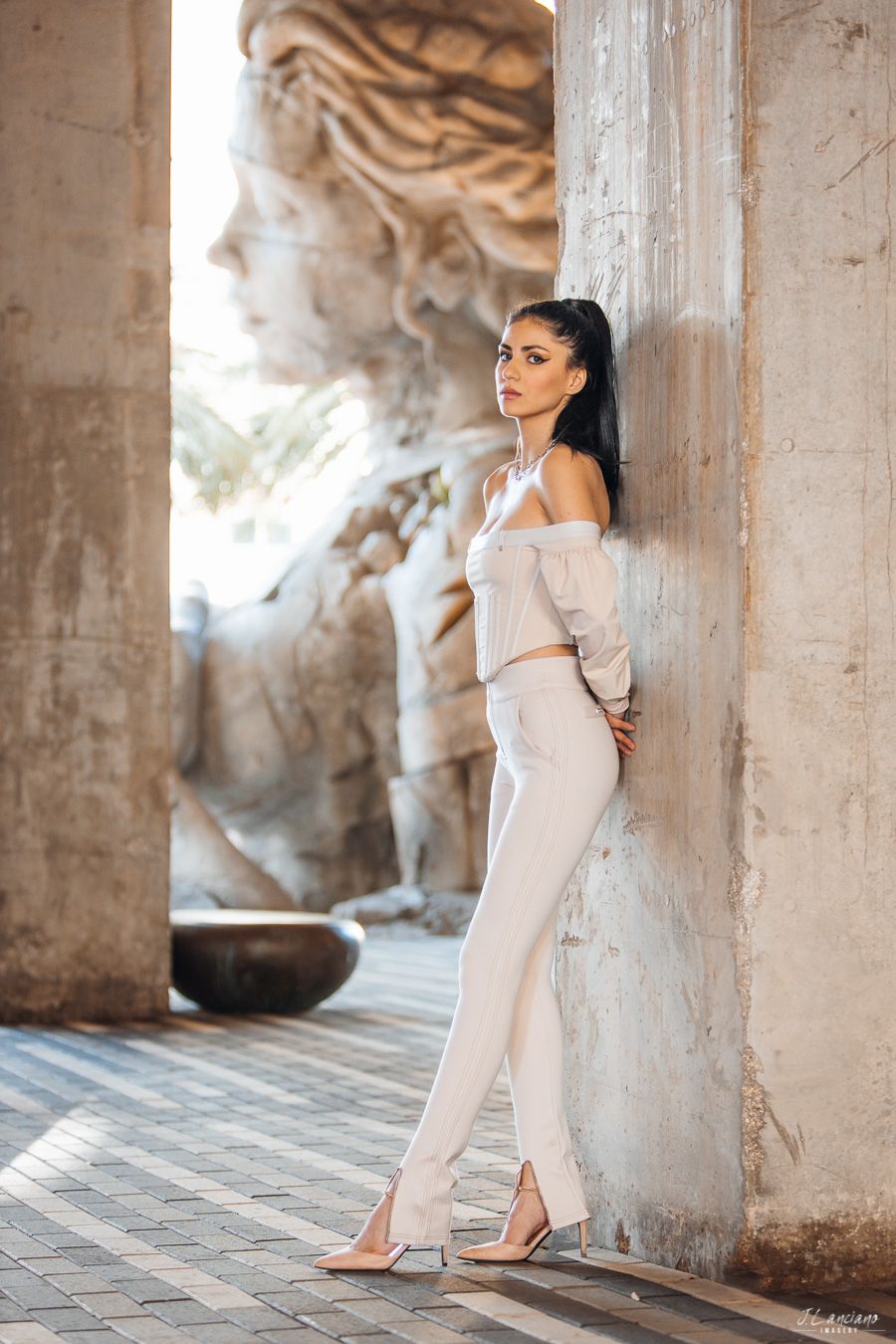 Woman in elegant cream outfit poses against stone column with classical relief sculpture.