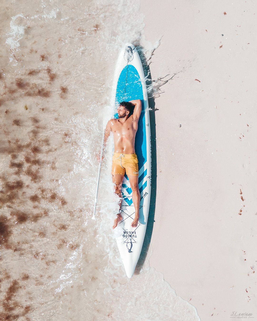 Aerial view of person relaxing on blue paddleboard on sandy beach.