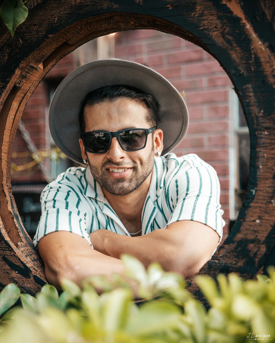 A person wearing sunglasses and a hat smiles through a wooden circular frame with greenery.