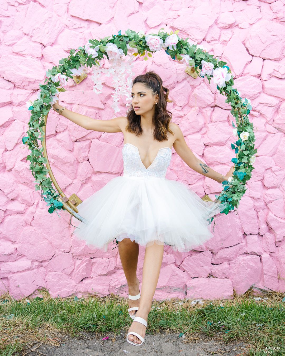 A dancer in a white tulle dress poses gracefully against a pink wall with a floral arch.