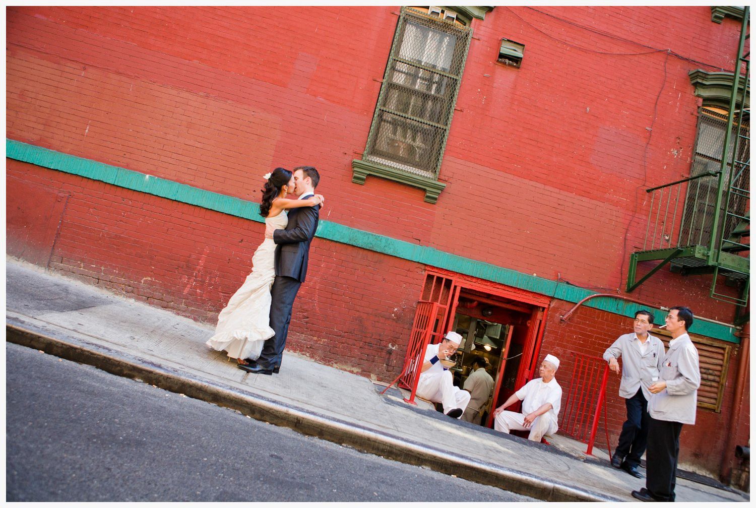 A couple embraces against a red brick wall while onlookers rest nearby on an urban street.