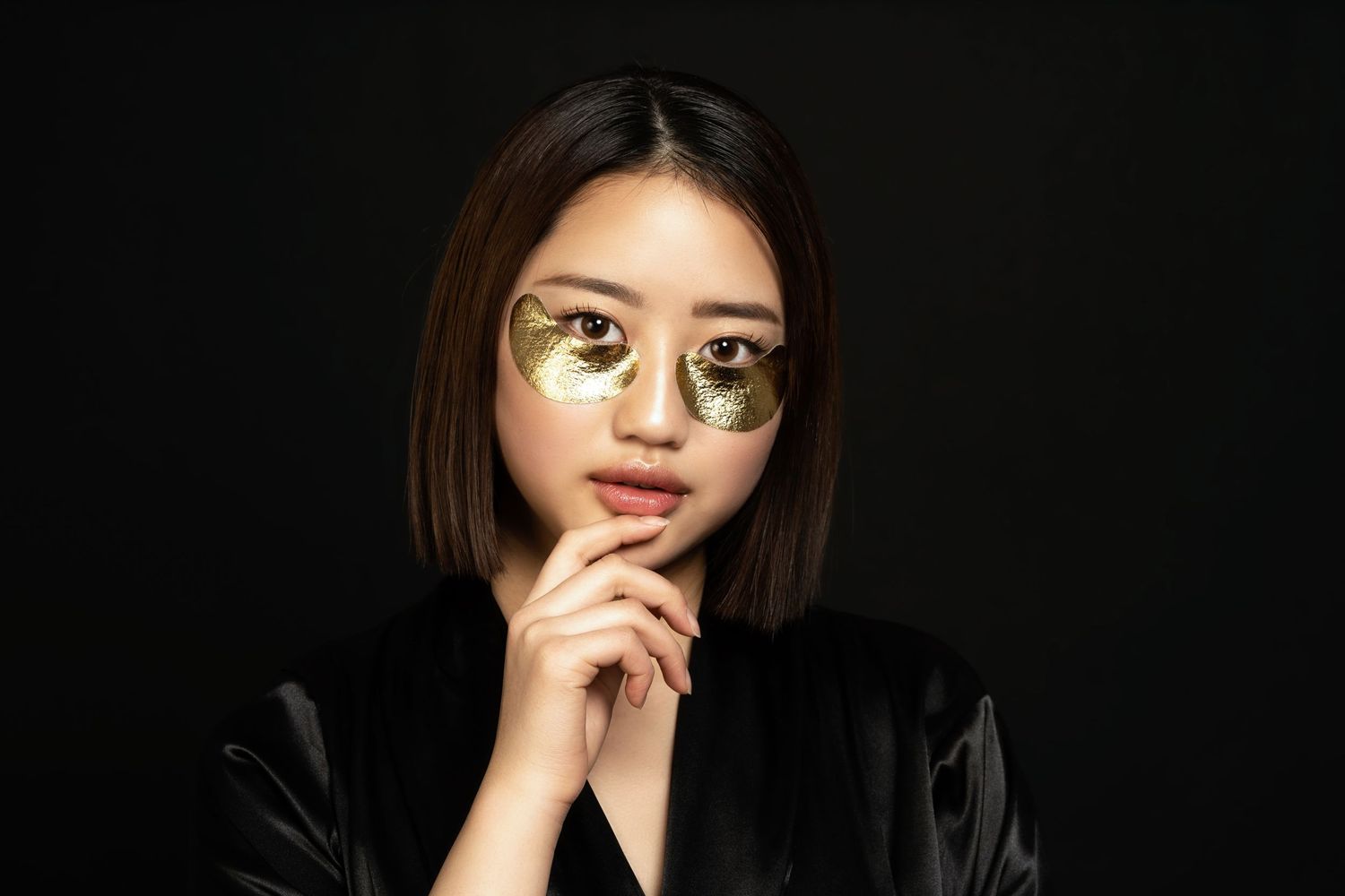 model fotografie voor een beauty merk, model heeft goudkleurige oog pads onder ogen en staat voor zwarte achtergrond, ze houdt kin vast.