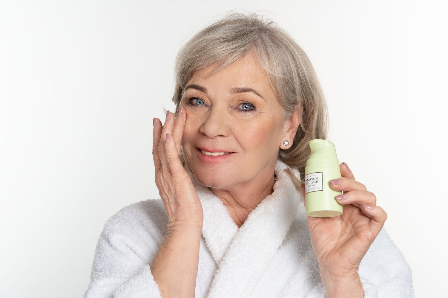 blond ouder model poseert met beauty product in linkerhand voor witte achtergrond en masseert creme op wang met rechterhand.
