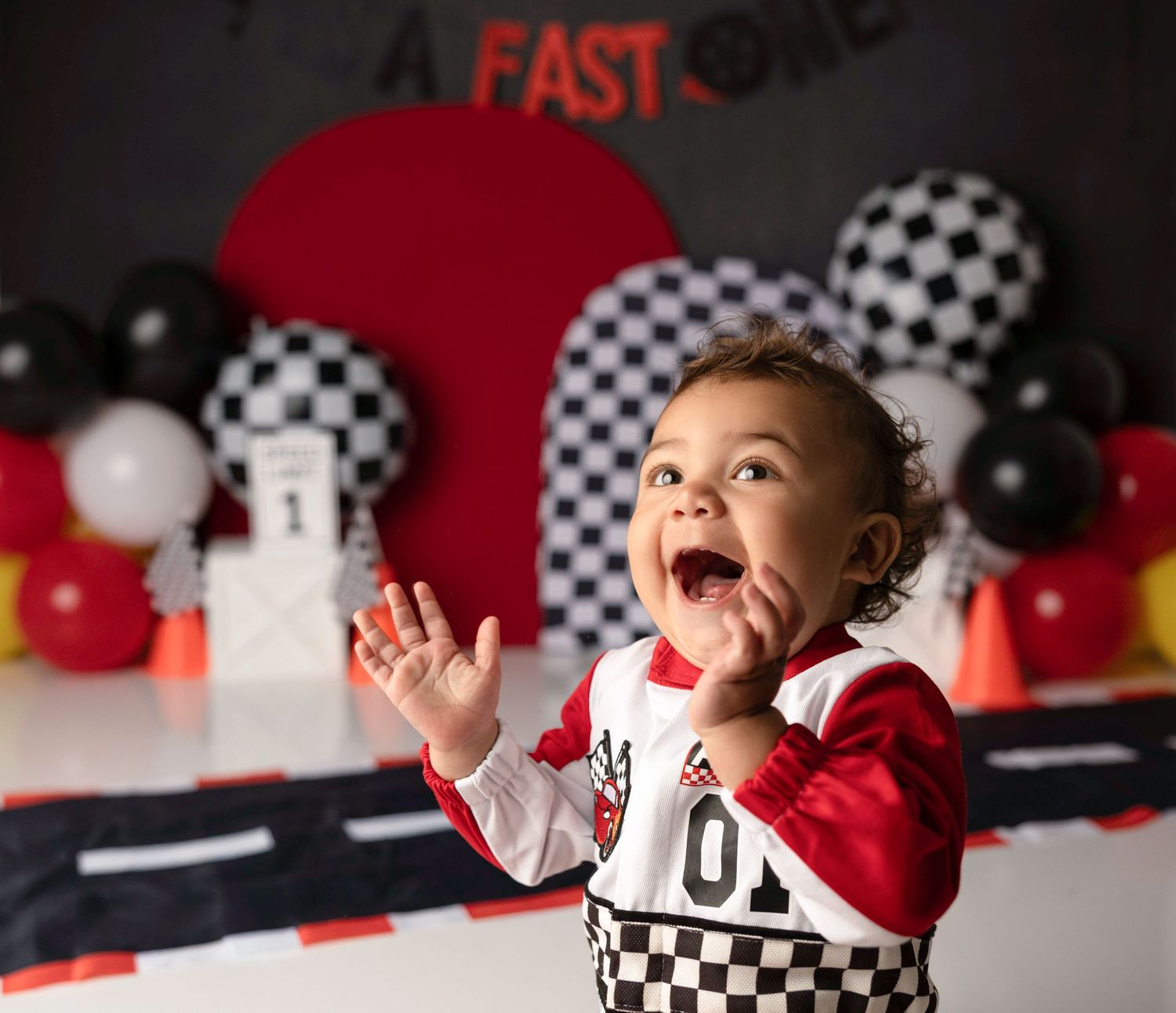Laughing baby in racing outfit claps hands at race car themed first birthday cake smash photo session.