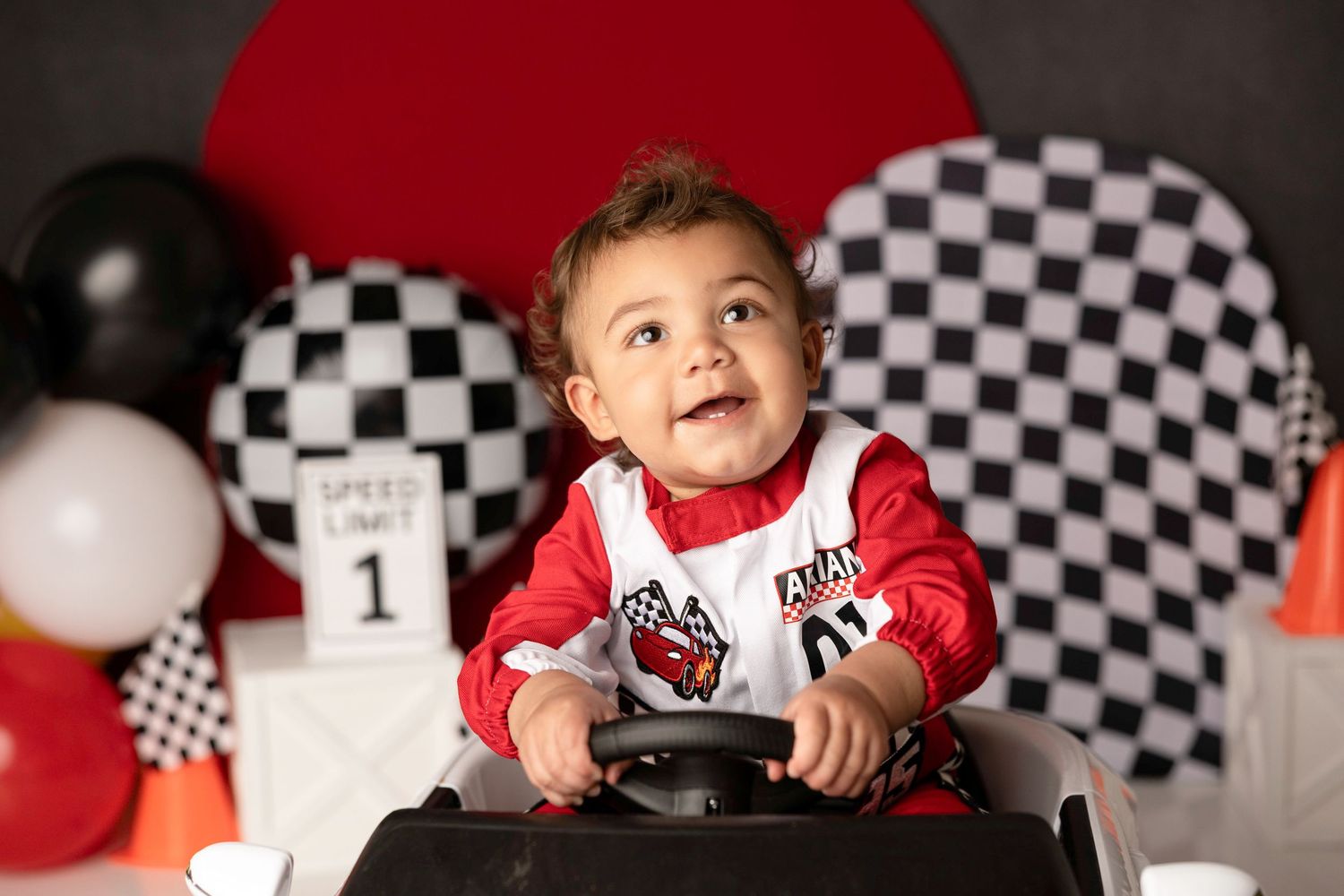 Baby boy in red racing outfit sits in toy car with checkered flags and balloons for his first birthday cake smash.