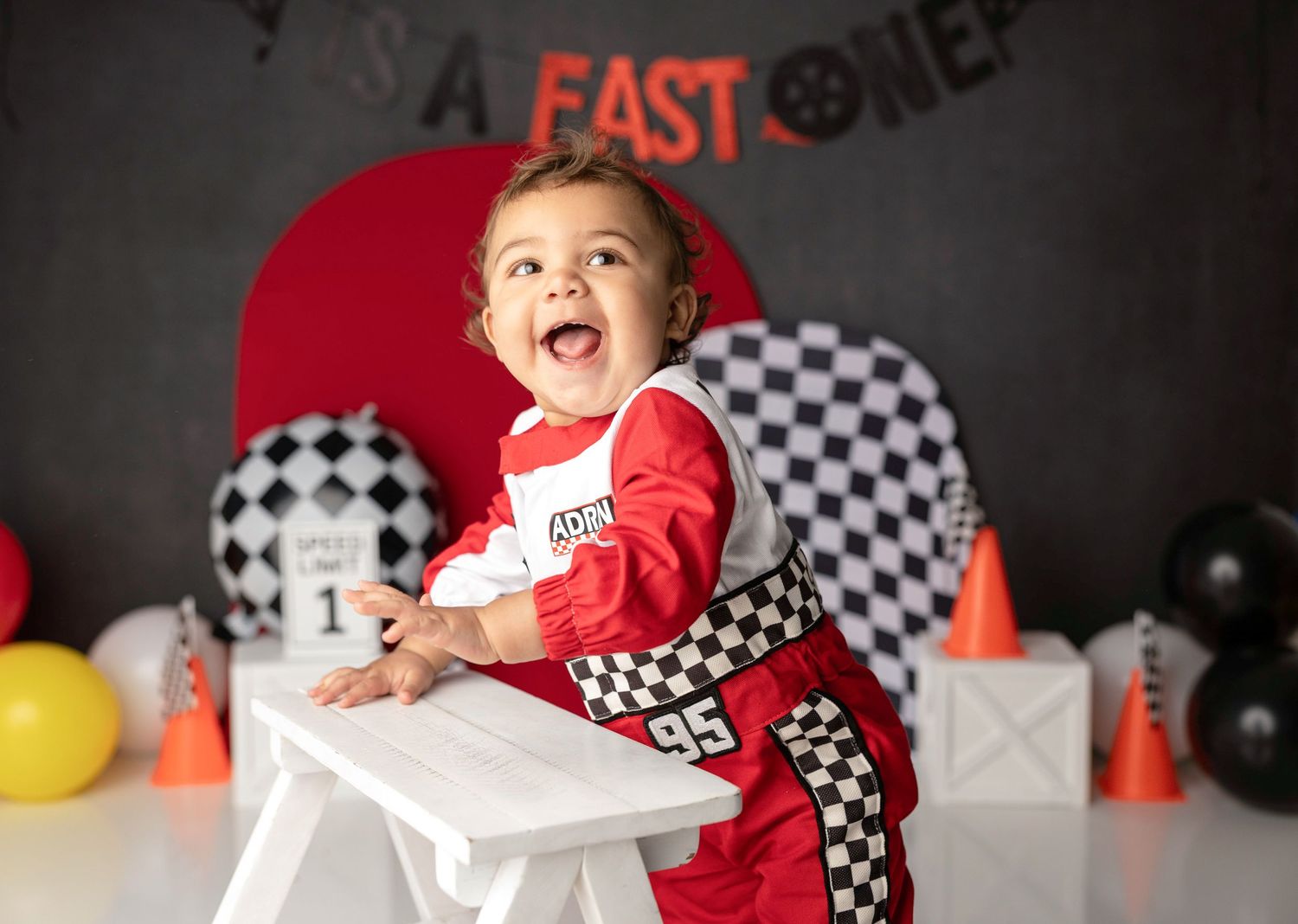 Happy baby in red checkered racing suit sits at white table at race car first birthday photo shoot.