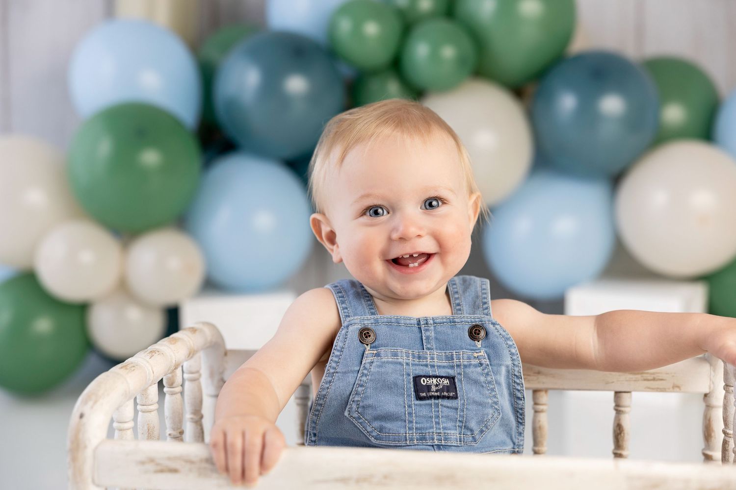 Happy baby boy in denim overalls smiling in front of green and blue balloon garland backdrop.