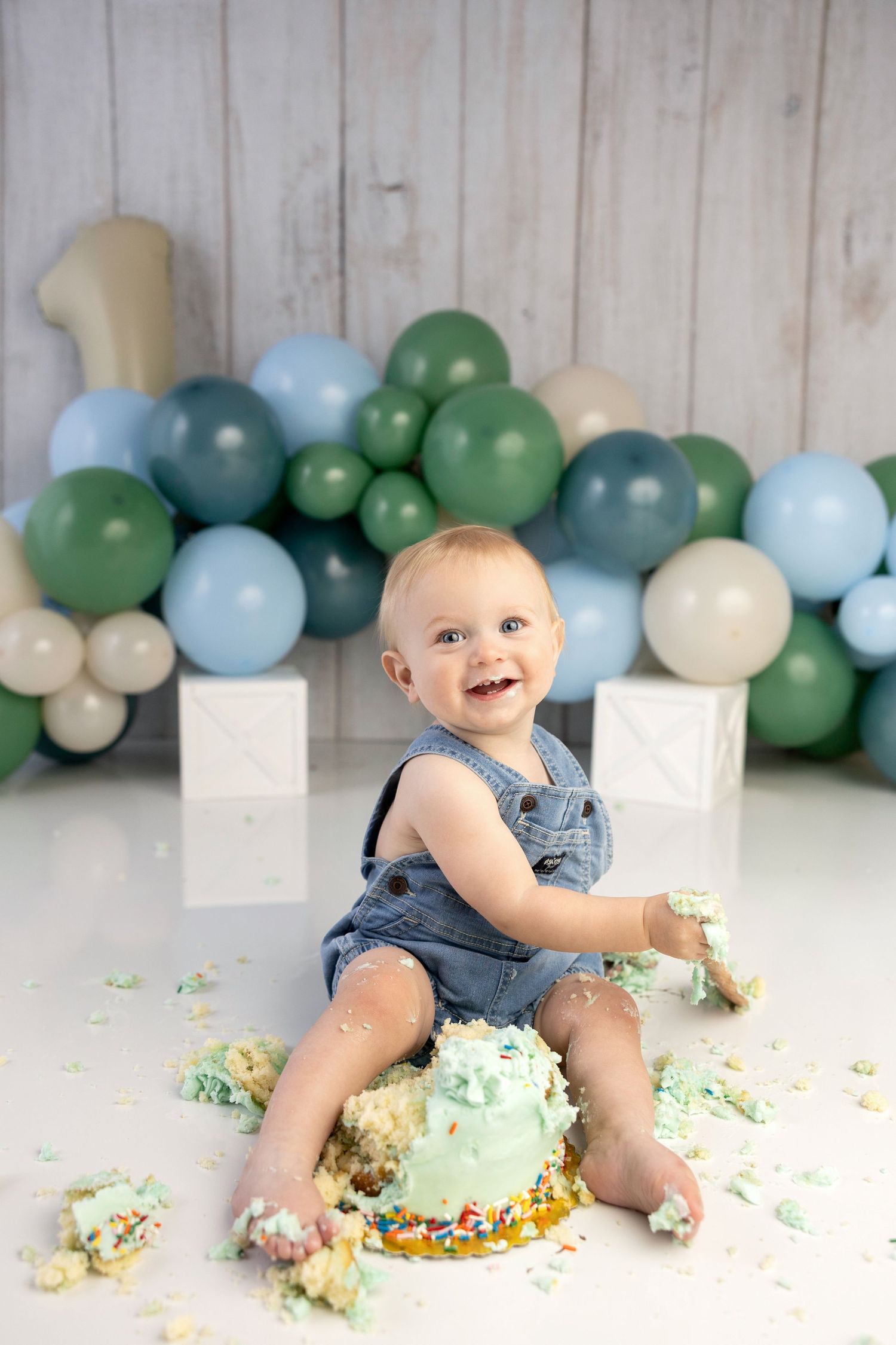 Smiling baby boy in denim overalls doing a cake smash at his first birthday with green and blue balloons.