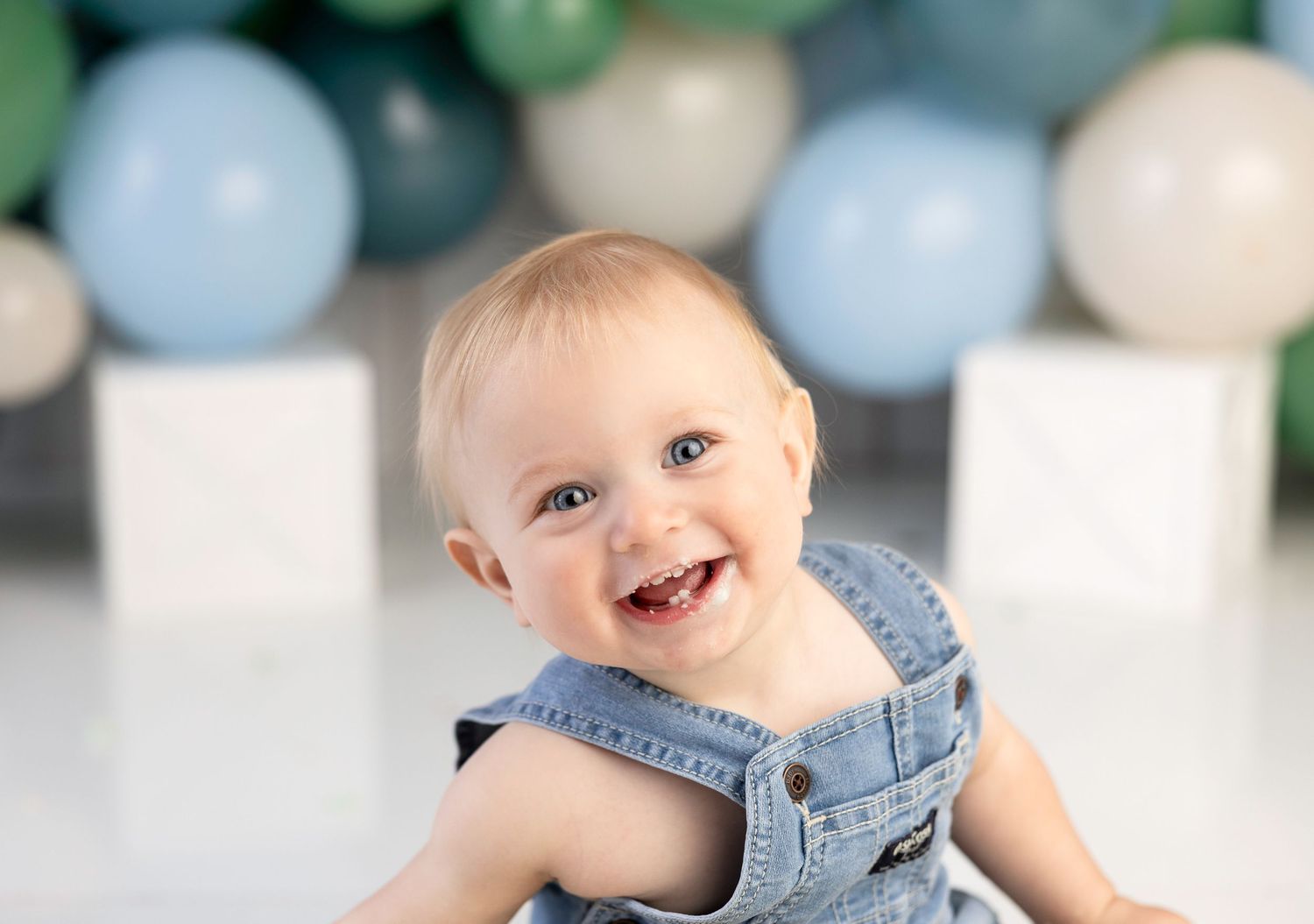 Adorable smiling baby in denim overalls sitting in front of blue and green balloon party backdrop.