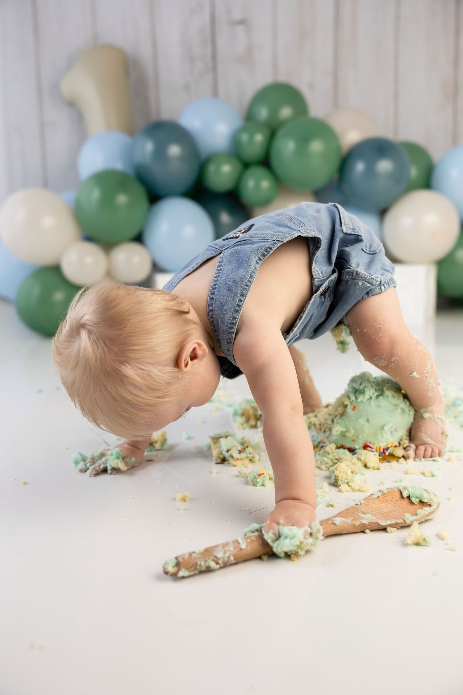 Toddler in denim overalls smashing first birthday cake surrounded by green and blue balloons.