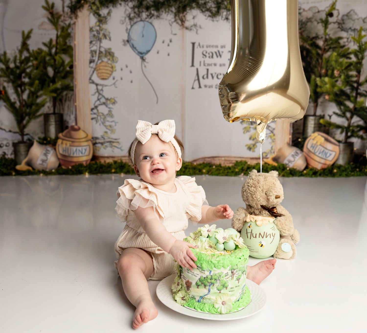Baby girl in cream outfit celebrates first birthday with green smash cake, teddy bear, and gold number one balloon.